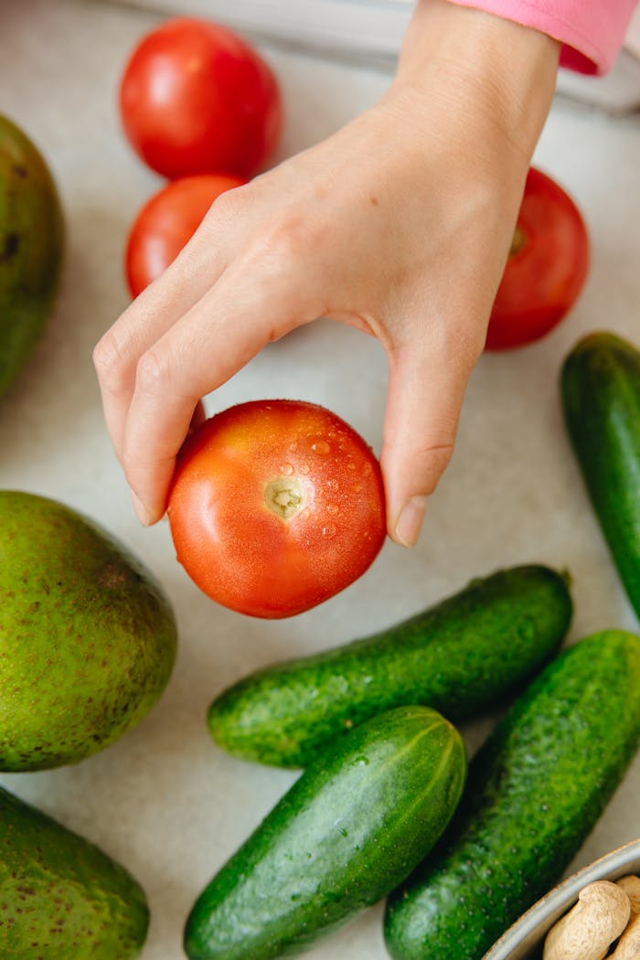Close-up of a hand picking a fresh tomato surrounded by cucumbers and avocados.