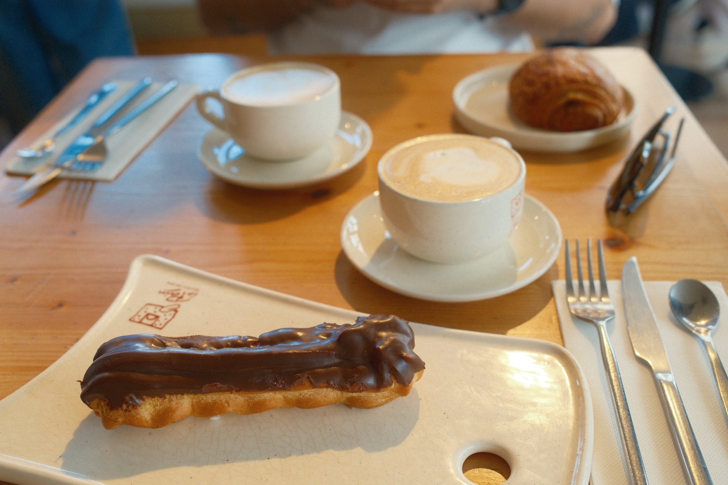Coffee and chocolate éclair on a wooden table in a cozy café. Breakfast setting with cappuccino, pastry, warm tones, lifestyle photography, café culture, morning routine, food photography.